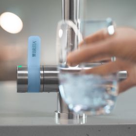 Frau hält vor der BRITA 3 Wege Küchenarmatur ein Wasserglas Eine Frau hält vor der BRITA 3 Wege Küchenarmatur ein halbvolles Wasserglas