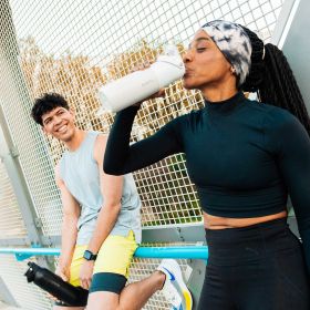 Zwei sportliche Personen machen eine Pause im Freien – eine trinkt aus einer weißen BRITA LARQ iQ Filterflasche, die andere hält eine schwarze Flasche und lächelt.